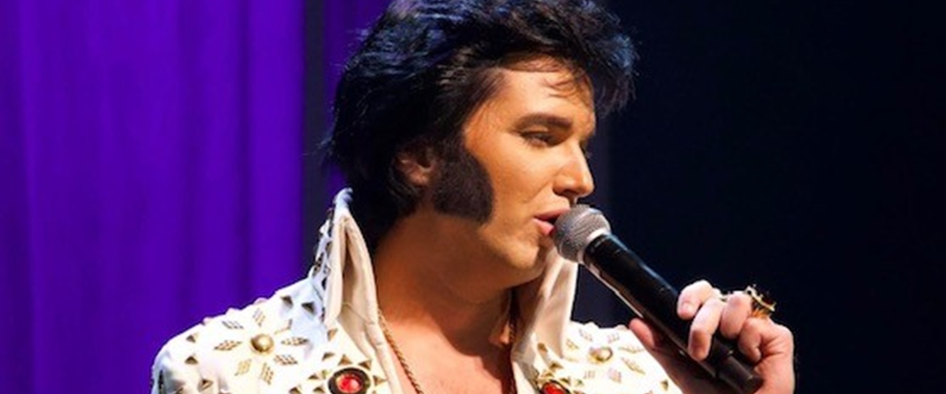 Elvis, Jimmy Holmes, forum Altoetting, Europolis, Bild Veranstalter Ein Mann in einem weißen Glitzeranzug hält ein Mikrofon in der Hand und singt.