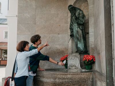Eine Stadtführerin steht mit einem Gast vor dem Bruder Konrad Brunnen, der sich gerade eine Flasche Wasser abfüllt. 