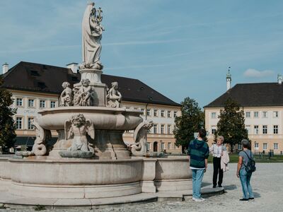 Eine Stadtführerin steht mit zwei Gästen vor dem Altöttinger Marienbrunnen. Stadtführung Herzerfrischendes Altötting, Foto Tourismusverband Inn-Salzach