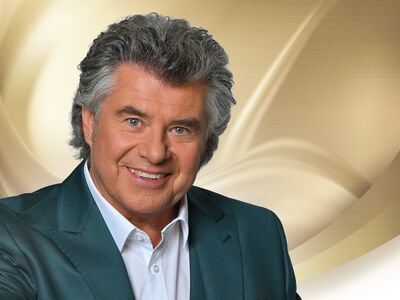 Ein Mann mit grau melierten Haaren und Anzug steht vor einem hellen Hintergrund. 