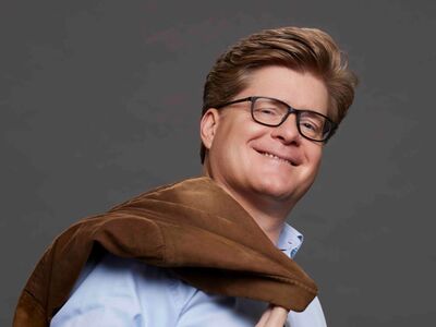 Herr Schröder, Forum Altötting, Bild Boris Breuer Der Kabarettist Johannes Schröder lacht mit Sakko über der Schulter in die Kamera.