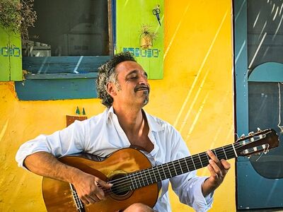 Trio Manuel Randi, Sud, Forum Altötting, Bild Veranstalter Ein Mann sitzt mit einer Gitarre vor eine bunten Hauswand.