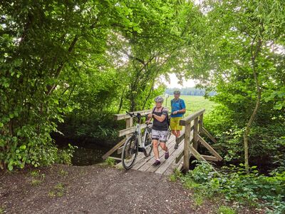 Geführte Radltour Altötting Erinnerungstour, Foto Dirschl Eine Dame und ein Herr schieben ihr Fahrrad über eine kleine Holzbrücke im Wald.