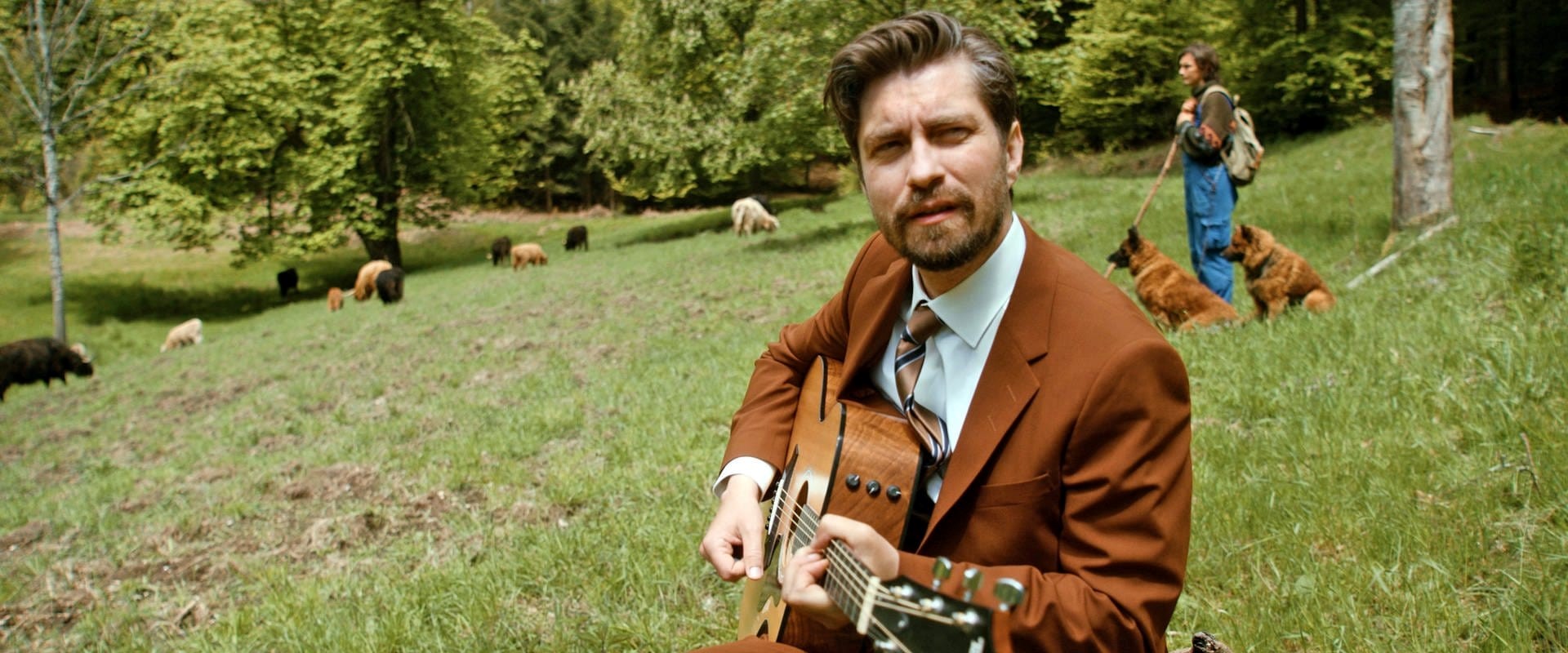 Der Musiker David Julian Kiurchner sitzt mit seiner Gitarre auf einer Schafwiese. 