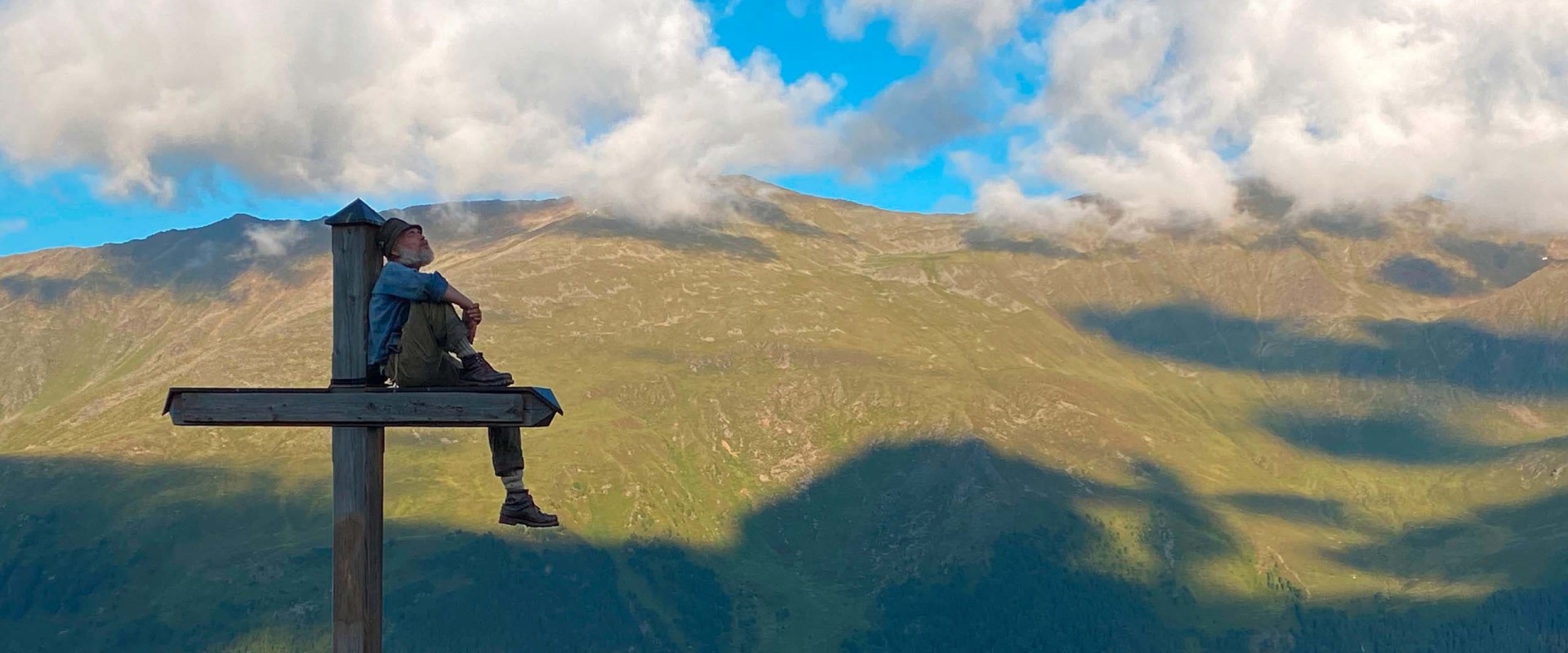 Der Aussteiger Eilias sitzt in der Bergwelt des Zillertals auf einem großen Holzkreuz.