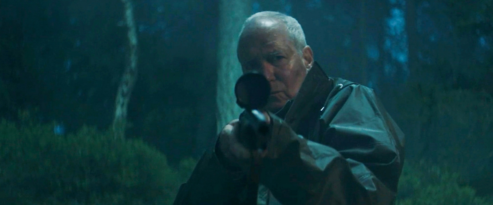 Der Schauspieler August Zirner hält eine Jagdwaffe direkt in die Kamera.
