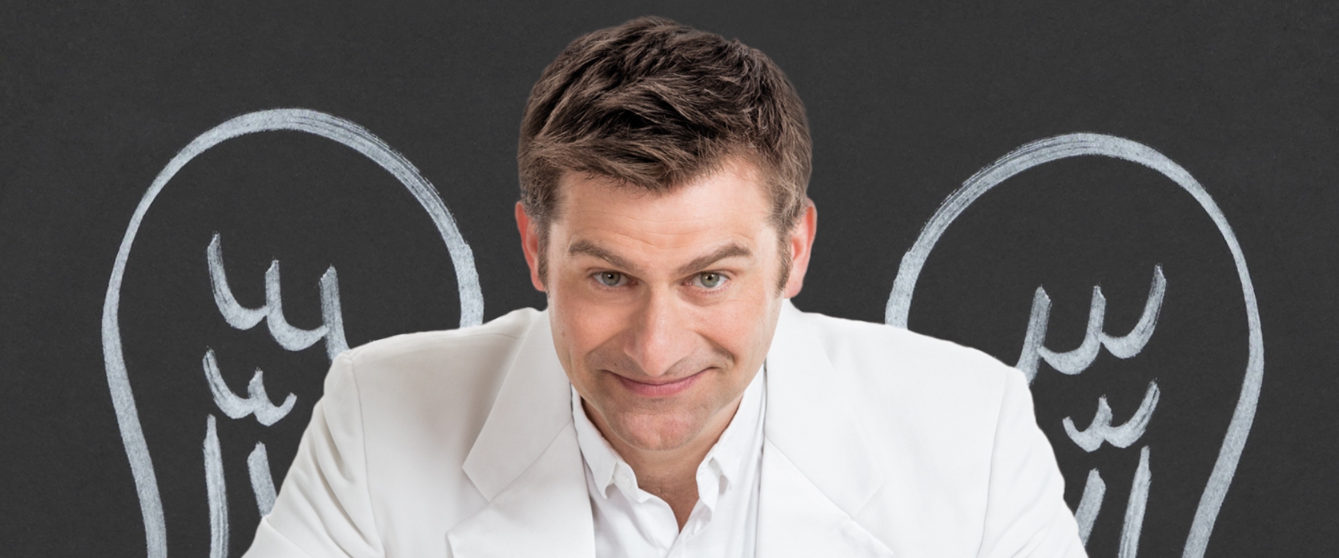 Michael Altinger sitzt mit Engelsflügel vor einem dunklen Hintergrund.