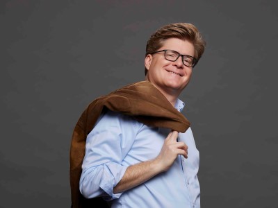 Herr Schröder, Forum Altötting, Bild Boris Breuer Der Kabarettist Johannes Schröder lacht mit Sakko über der Schulter in die Kamera.