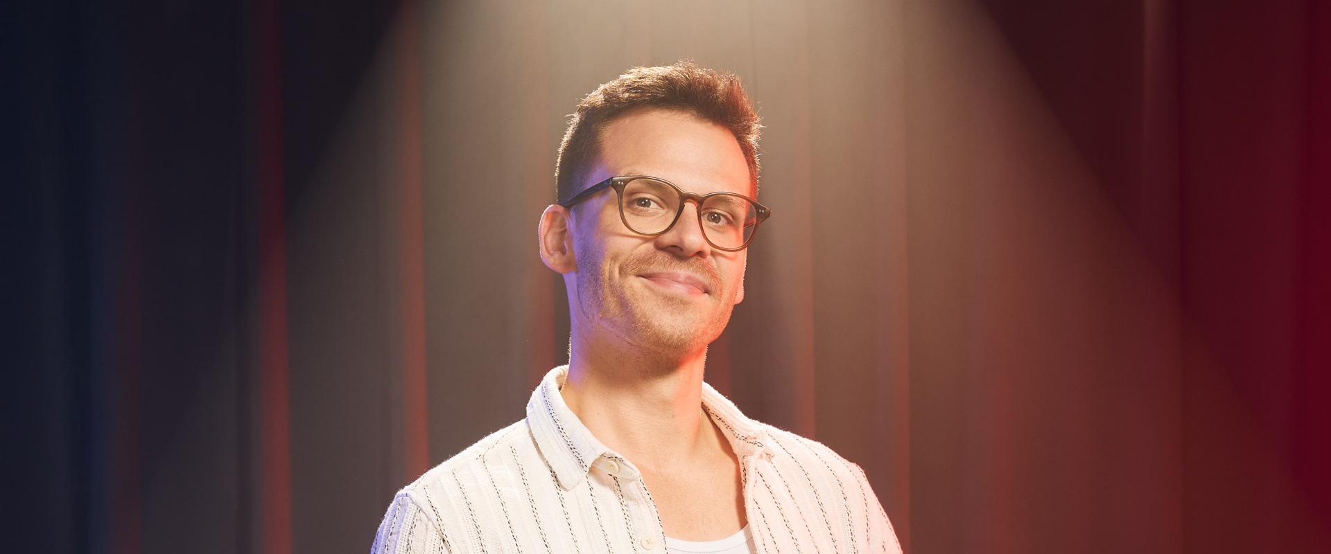 Ein Mann mit kurzärmeligem Hemd steht vor einem roten Vorhang.
