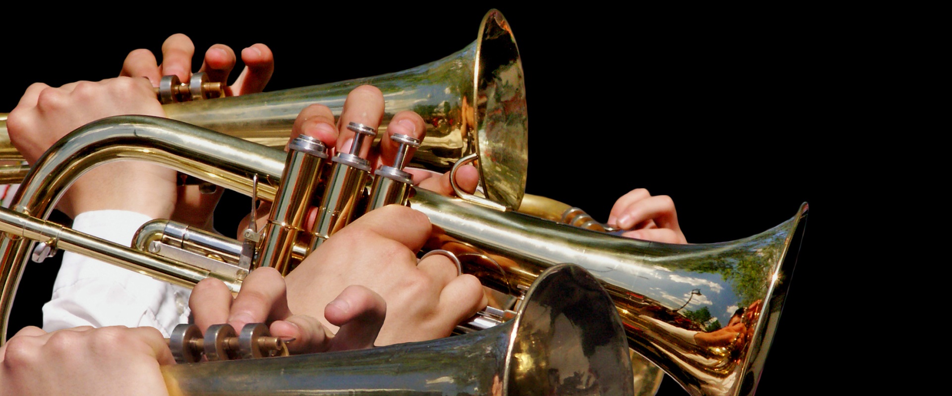 Es sind vier Trompeten mit acht Händen vor schwarzem Hintergrund zu sehen.
