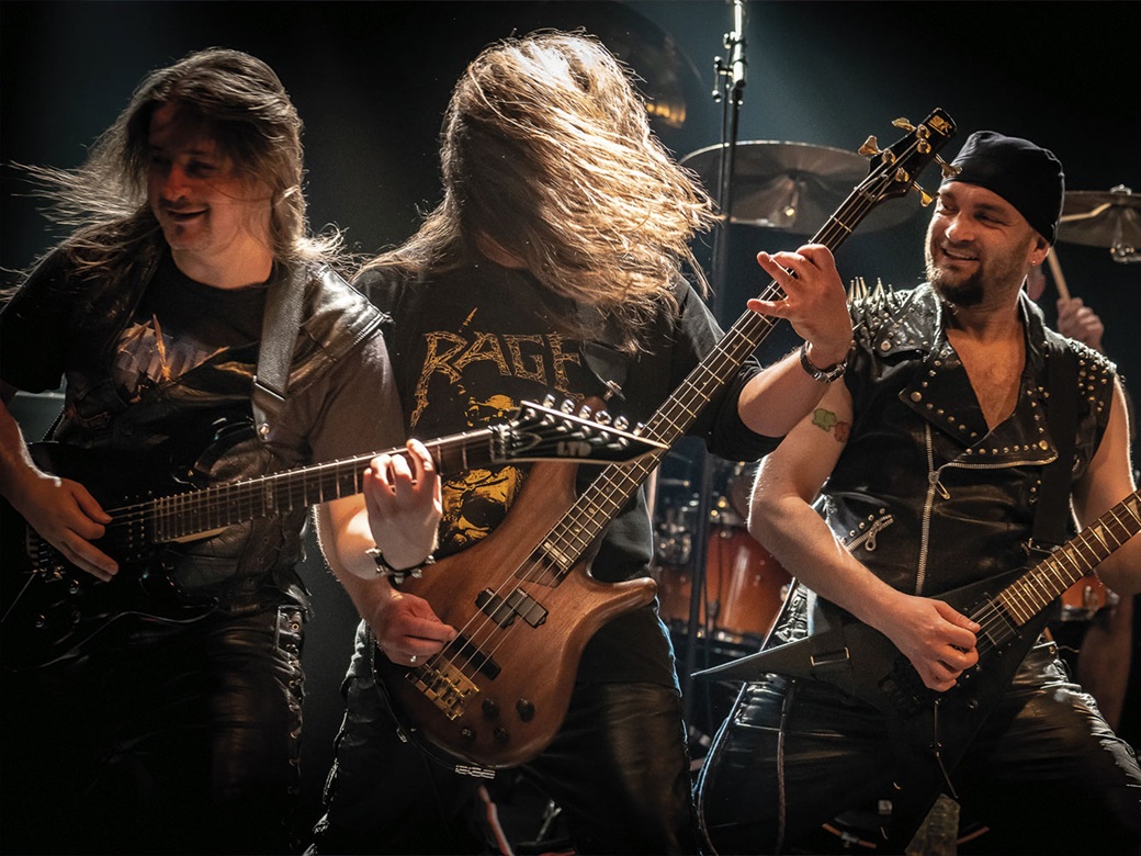 Drei Männer mit Gitarren stehen auf der Bühne und schwingen die Haare zum Head Banging.