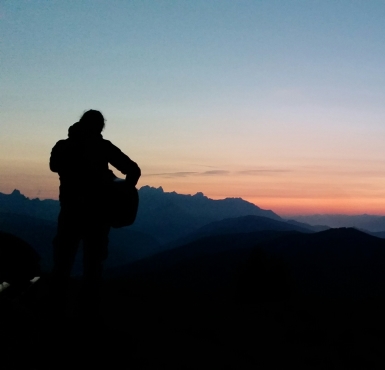 Alfons Hasenknopf steht auf einem Berg mit einer Gitarre und sieht in den Sonnenuntergang.