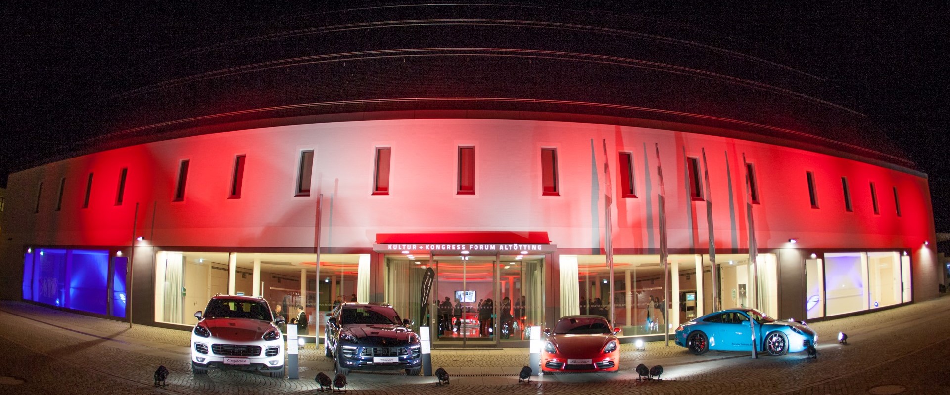 Eine Außenaufnahme des Altöttinger Forums bei Nacht, rot beleuchtet und mit verschiedenen Porsche Auto´s im Vordergrund.