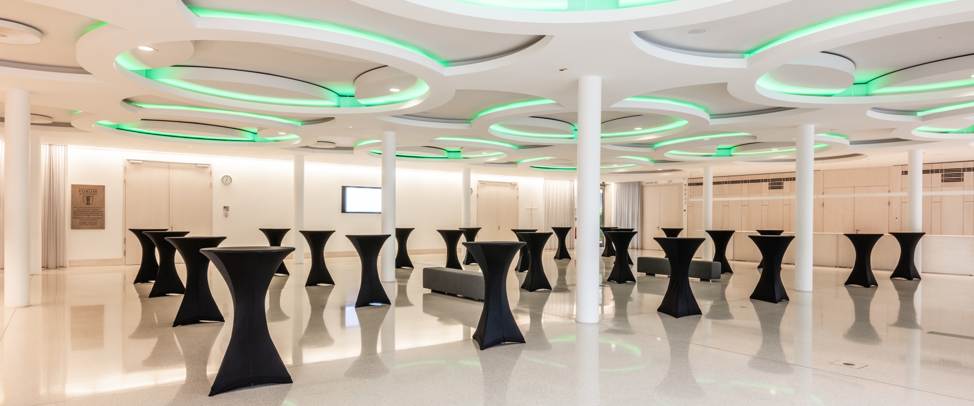 Das Foyer Abends im Forum Altötting, mit Stehtischen, die schwarze Hussen tragen und grüner Beleuchtung.